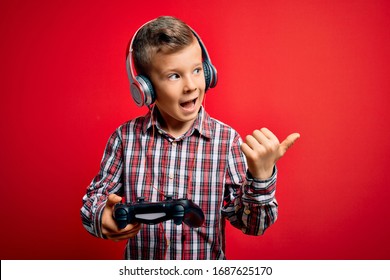 Young Little Caucasian Gamer Kid Playing Video Games Using Gamepad Joystick Over Red Background Pointing And Showing With Thumb Up To The Side With Happy Face Smiling