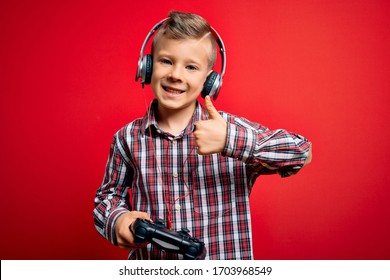 Young Little Caucasian Gamer Kid Playing Video Games Using Gamepad Joystick Over Red Background Happy With Big Smile Doing Ok Sign, Thumb Up With Fingers, Excellent Sign