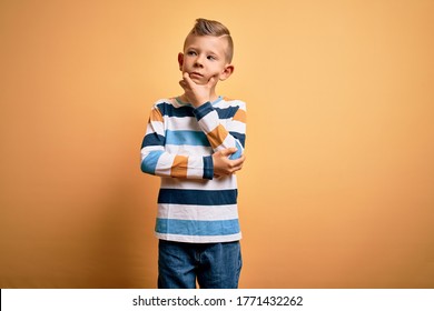 Young little caucasian kid with blue eyes wearing colorful striped shirt over yellow background with hand on chin thinking about question, pensive expression. Smiling with thoughtful face. Doubt.