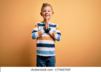 Young little caucasian kid with blue eyes wearing colorful striped shirt over yellow background praying with hands together asking for forgiveness smiling confident.