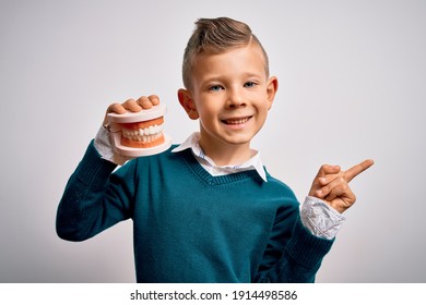 Young little caucasian kid holding dental prosthesis teeth denture over isolated background very happy pointing with hand and finger to the side
