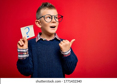 Young little caucasian kid wearing glasses holding paper note with question mark sign pointing and showing with thumb up to the side with happy face smiling