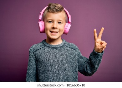 Young Little Caucasian Kid Wearing Headphones Listening To Music Over Purple Background Smiling With Happy Face Winking At The Camera Doing Victory Sign. Number Two.