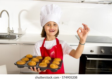 Young Little And Cute Girl Alone In Cook Hat And Apron Presenting And Showing Tray With Muffins Smiling Happy And Proud In Learning Baking Concept At Modern Home Kitchen