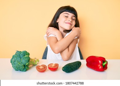 Young Little Girl With Bang Sitting On The Table With Veggies Hugging Oneself Happy And Positive, Smiling Confident. Self Love And Self Care 