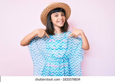 Young little girl with bang wearing summer dress and hat looking confident with smile on face, pointing oneself with fingers proud and happy. 