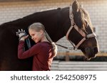 Young little teenager kid girl portrait hugging chestnut horse  at stable ranch yard. Horse riding school exercise. Cute little beginner blond girl near beautiful brown pet. Equine hobby therapy