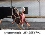 Young little teenager kid girl leading chestnut horse through stable ranch yard. Horse riding school exercise.  Cute little beginner blond girl kid near beautiful brown horse. Equine hobby or work