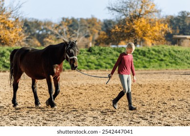 Young little teenager kid girl leading chestnut horse through stable ranch yard. Horse riding school exercise.  Cute little beginner blond girl kid near beautiful brown horse. Equine hobby or work - Powered by Shutterstock