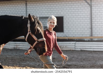 Young little teenager kid girl leading chestnut horse through stable ranch yard. Horse riding school exercise.  Cute little beginner blond girl kid near beautiful brown horse. Equine hobby or work - Powered by Shutterstock