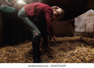 Young little teenager kid girl cleaning horseshoe hoof picking brush tools chestnut horse at stable ranch. Horse ride school farm life. Cute little blond girl kid  care brown horse. Equine hobby work - Powered by Shutterstock