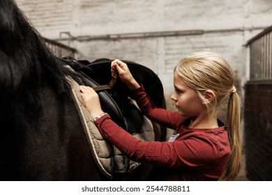 Young little teenager kid girl adjusting saddle chestnut horse back farm stable ranch preparing training. Horse ride school farm life. Cute little blond girl kid care brown horse. Equine hobby work - Powered by Shutterstock