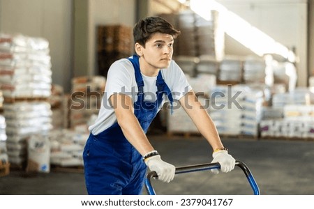 Young loader guy in overalls rolls cargo cart in warehouse