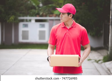 Young Logistic Career Concept. Happy Delivery Man Looking For His Customers At Their Home. Taken In Real House. Asian Chinese Fit Man In Red Polo Shirt And Jeans With Red Hat In His Early Twenties.