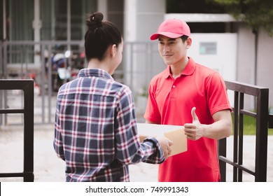 Young Logistic Career Concept. Happy Delivery Man Giving His Package To Customer At Home. Taken In Real House. Asian Chinese Fit Man In Red Polo Shirt And Jeans With Red Hat In His Early Twenties.