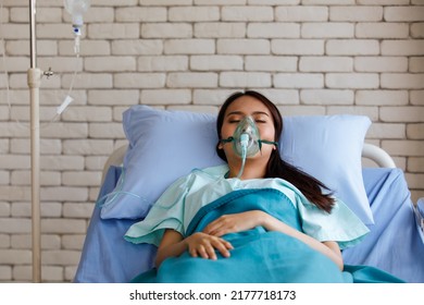 Young Long Black Hair Sick Asian Female Patient Wear Green Shirt And Oxygen Intubation Mask On Face Sleeping On Blue Sheet Hospital Pillow And Bed In Icu Ward Room With White Brick Wall Background.