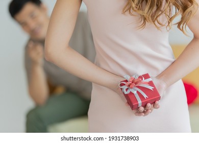 A young long brown hair female wearing pink sleeveless dress stand hiding red gift box with white ribbon behind her back when her boyfriend sitting on orange couch waiting for surprise.
