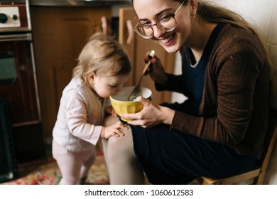 Young Long Haired Beautiful Mother Preparing Food In Kitchen For Her Little Funny Daughter. Indoor Lovely Girl Portrait. Parent With Child Making Dinner At Home. Happy Family Together. Room Mess