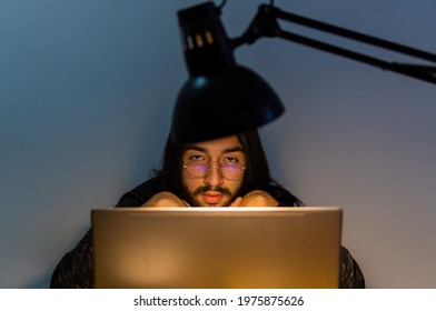 Young Long Haired Latin Man With Glasses Using Laptop Computer, Looking Straight At Camera Over Glasses.architect's Lamp, Metal Swing Arm Lamp