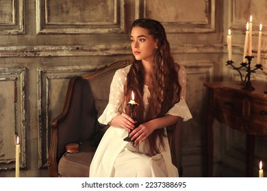 Young long haired woman in vintage night gown sitting on chair with candle in hands. Historic portrait of noble woman