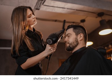 Young long long-haired female hairdresser hairstyling drying hair with professional hair dryer bearded man in Salon Chair. Modern black-style barber shop.Hair Care service local small business concept - Powered by Shutterstock