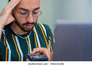 Young, Long-haired Latino Man With A Worried Look On His Face, Doing Maths On A Calculator, While Using The Computer.