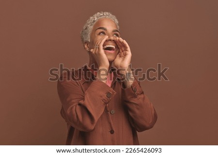 Young loud horny African American woman in casual clothes puts hands to mouth to shout loudly about beginning of discount week at mall or invite to house party stands on brown background
