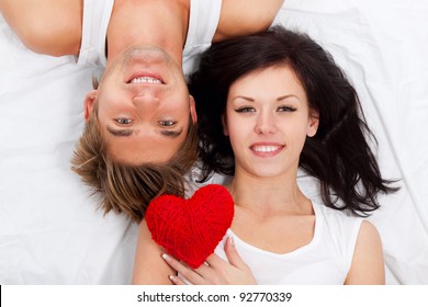 Young Lovely Couple Holding Red Heart Together Lying In A Bed, Happy Smile Looking At Camera, Valentine Day Love Concept