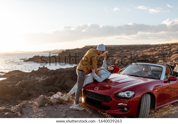 Young lovely couple kissing on the beach,\
traveling by car on the rocky ocean coast on a sunset. Carefree\
lifestyle, love and travel\
concept