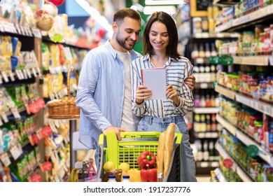 Young Lovely Couple With Shopping Cart Full Of Healthy Eco Products Looking Into Their Grocery List At Supermarket. Smiling Man Embaricng Woman While Buying Products At Mall Together