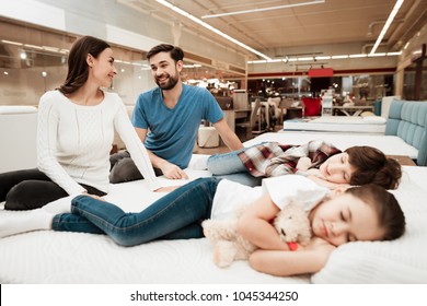 Young lovely couple is sitting on mattress next to sleeping children in furniture store. Testing softness of mattress. Choosing mattress in store