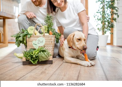 Young Lovely Couple Sitting Together With Their Dog And Fresh Green Vegetables At Home