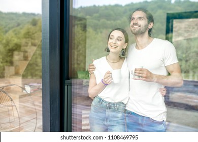 Young And Lovely Couple Standing Together Looking Outside The Window At Home, View Through The Window Vith Reflection