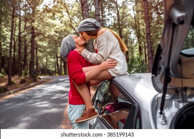 Young and lovely couple in sweates and hats having fun hugging together near the car on the roadside in the pine forest