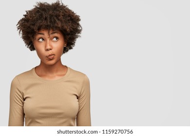 Young lovely female has dark skin, clueless and unaware expression, purses lips as being questionned, looks with puzzlement upwards, isolated over white background with copy space on right side