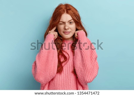 Young lovely ginger female musician plugs ears as hears awful noise or music, squints face, has long wavy foxy hair, wears oversized pink jumper, avoids loud sound, ignores something unpleasant