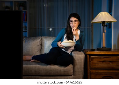 young lovely girl holding popcorn box and looking at tv screen enjoying movie showing puzzled emoticon sitting in living room comfortable sofa eating snack at night.