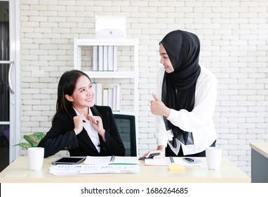 Young Lovely Muslim Businesswomen Happy And Give A Thumbs Up To Young Pretty Lady Buddhist In Office Room And Talking Discussion About Success In Online Business Job. Bangkok Thailand South East Asia