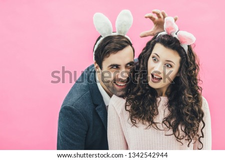 Young lovers couple on the pink background. With a bunny ears on the head. Surprised, the wife opened her big eyes, while the man held his hand over his head. Easter.