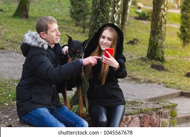 A Young Lovers Couple Sits In A Spring Park With Their Dog And Use The Phone