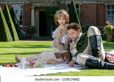 Young lovers dressed in vintage clothing sitting on picnic blanket. Gentleman is reading to his lover from a book of poems