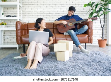 Young lovers spend time together on holidays in the living room. Asian man with headphones playing an acoustic guitar while the girl check the parcel delivery schedule by laptop computer. - Shutterstock ID 1875117148