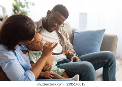 Young Loving African American Parents Feeding Their Baby Boy From Bottle, Cute Black Toddler Drinking Water Or Milk. Millennial Family Sitting On Couch, Spending Time With Little Child