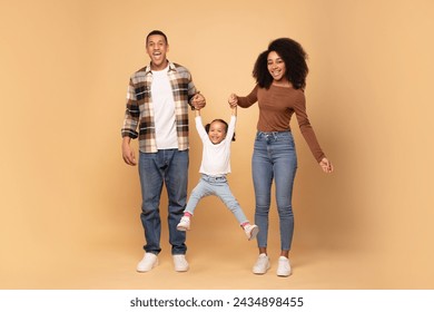 Young loving black parents playing with daughter, holding hands and girl jumping, family posing over beige studio background, full length shot - Powered by Shutterstock