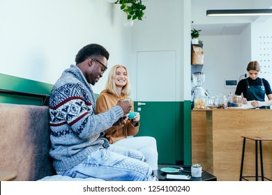 Young Loving Couple Blond Woman And African American Man In Warm Sweater Sitting With Mint Green Coffee Cups In The Modern Minimalism Interior Cafe In Winter Time With Barista On Background.