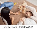 Young loving couple, happy family owners playing with cute pet puppy at home, spouses enjoy time together having fun with small pedigreed dog on carpet, indoor leisure activity in new living room