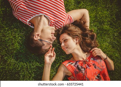 Young Loving Couple Lying Together Head To Head On A Grass At Summer. Family Picnic. Both In Red Clothes And Holding Hands. Overhead Top View.