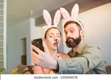 Young Loving Couple Making Selfie On Mobile Phone With Pink Rabbit Ears On Head. Happy Family Preparing For Easter