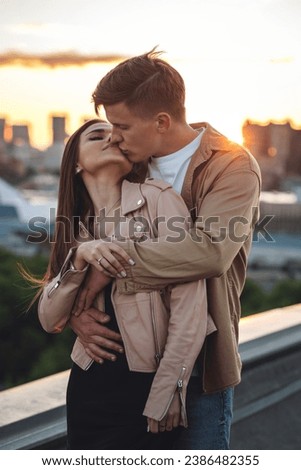 Young loving couple on a surprise date on rooftop on Saint Valentine's Day. Laughing, kissing embracing, drinking wine, having candlelit picnic with cityscape urban view with skyscrapers on background