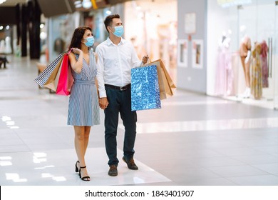 Young Loving Couple In Protective Medical Mask With Shopping Bags Walking In The Mall. Shopping During The Coronavirus Epidemic. Sale, Consumerism, Black Friday Concept. 